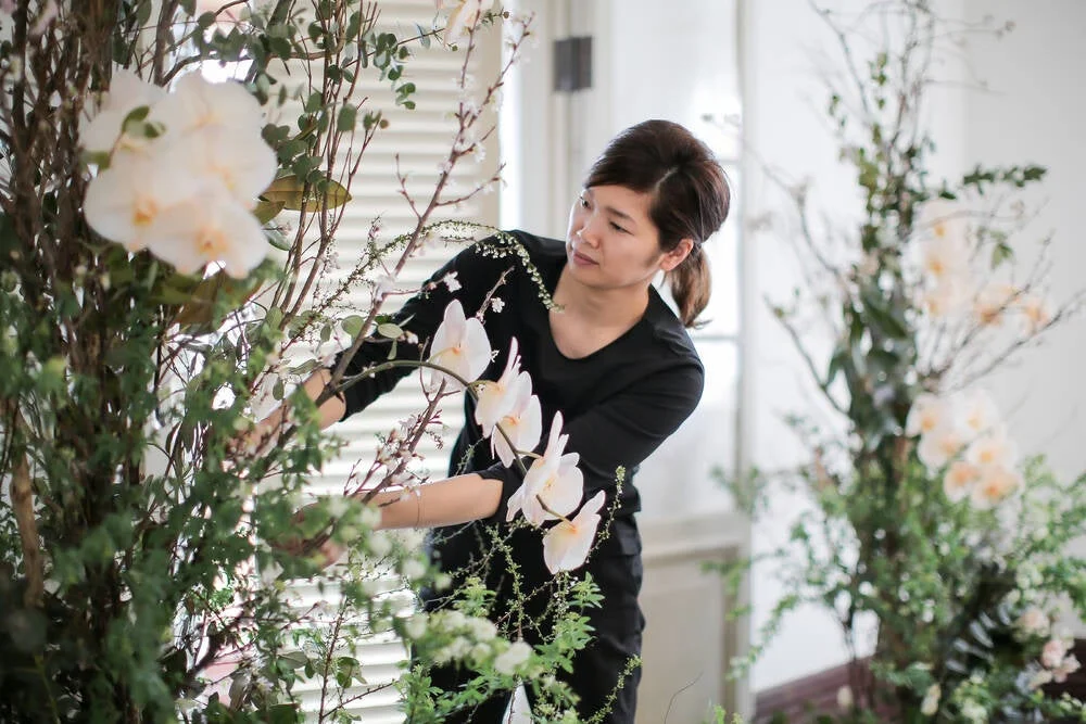 女性が白い胡蝶蘭や緑の植物を使って大きなフラワーアレンジメントを制作している様子です。エレガントな空間での装飾作業で、集中して花と向き合っています。